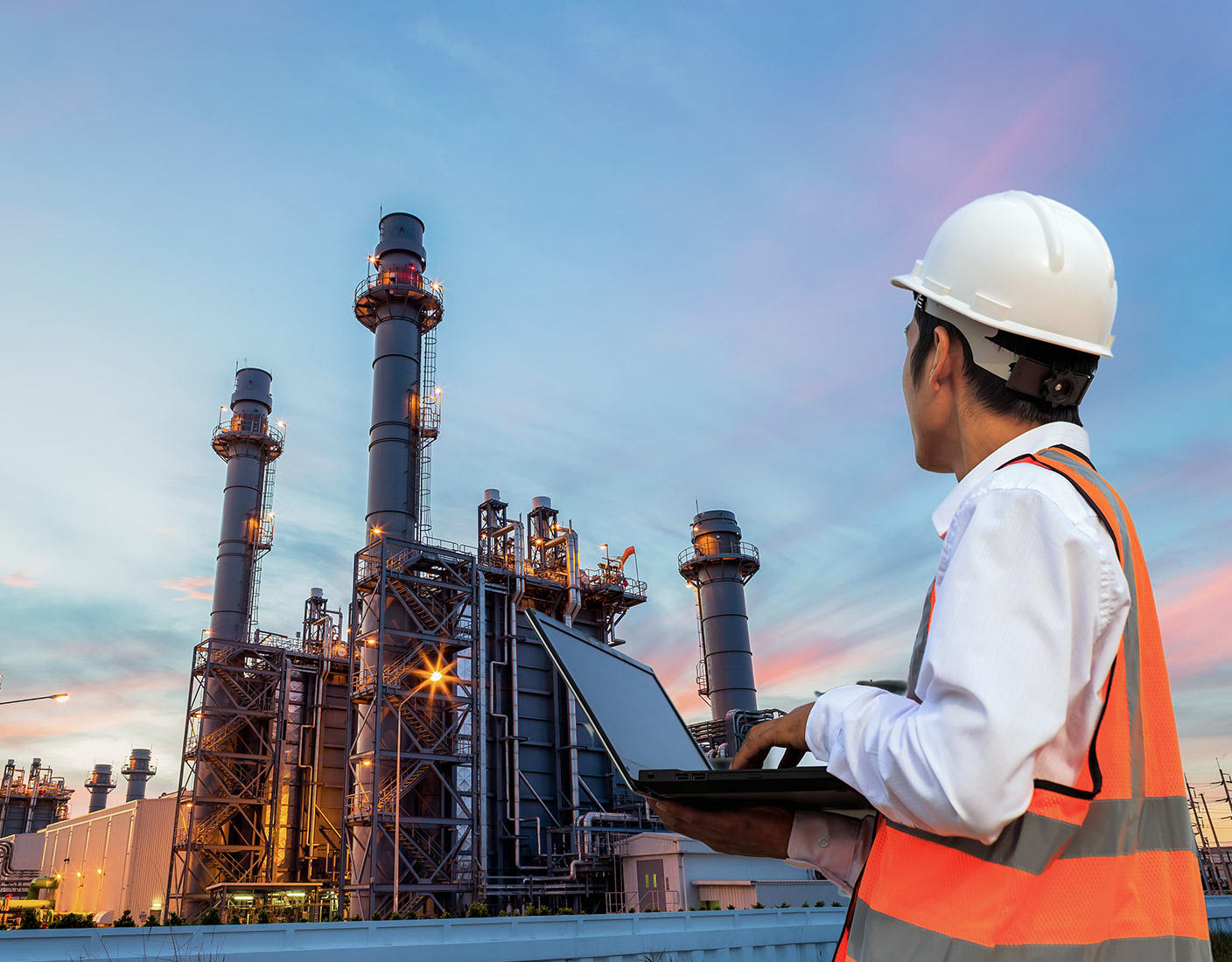 A worker in a white hard hat and orange safety vest inspects an industrial plant while using a laptop. The plant's chimneys and infrastructure are visible under a cloudy sky at dusk.